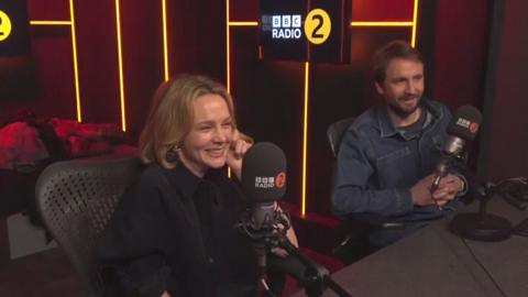 A woman with blonde hair and black top sits smiling in a Radio 2 studio. A man in a denim shirt sits beside her