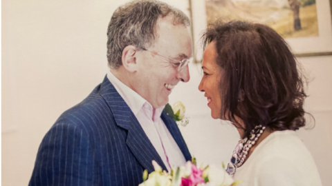 Andrew and his wife Zoubida on their wedding day. He is in a navy striped suit, she is in a white dress and bold blue and silver necklace, they are looking at one another and smiling. 