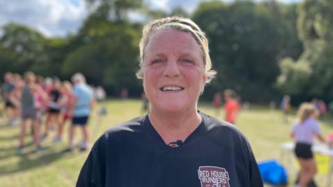 Hayley Myford is standing in Sutton Park wearing running clothes, including a black t-shirt and a head band. She is smiling at the camera. Behind her you can see other runners.