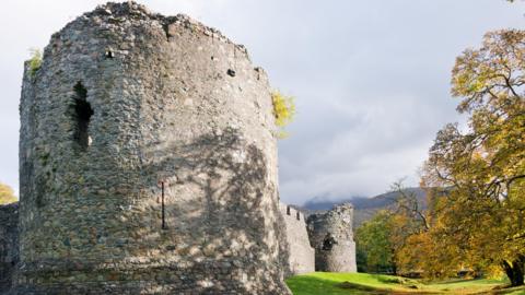 The image show a corner tower at Old Inverlochy castle. Shadows from trees are being cast on to the stonework. The picture shows an autumnal scene with leaves turning yellow and orange. further parts of the castle's wall and another corner tower can be seen in the background along with mist covered mountains. 