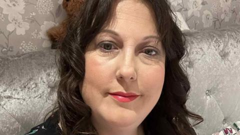 A woman with brown hair looks into the camera whilst sat on a grey sofa.