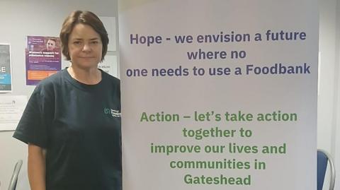 Diane Kindred standing next to a Gateshead Foodbank banner. She has dark hair and is wearing a dark blue T-shirt. The banner includes a number of action points such as "Hope - we envision a future where no one needs to use a foodbank".