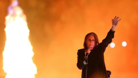 Image shows Ozzy Osbourne on stage in Birmingham at the closing ceremony of the Commonwealth Games. The background of the picture is orange and to the right is a large flame. Ozzy is on the left with his trademark long dark hair, black clothes and black eyeliner around his eyes. He is holding the microphone with his right hand and his left hand is in the air. 