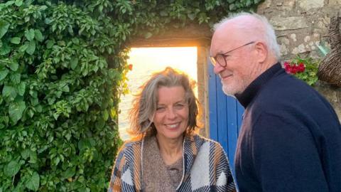 Sue Butler, with blue eyes and grey hair and a loose white, blue and orange poncho style top, with her husband Eddie Butler, a tall man with grey hair and glasses and a navy blue turtle neck sweater. They're stood in front of a stone house with an open blue door and a wall of green leaves 