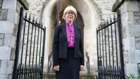 Dame Sarah Mullally stands in front of grey stone building in the middle of half opened black gates. She wears a black suit with a purple shirt, dog collar and cross around her neck.