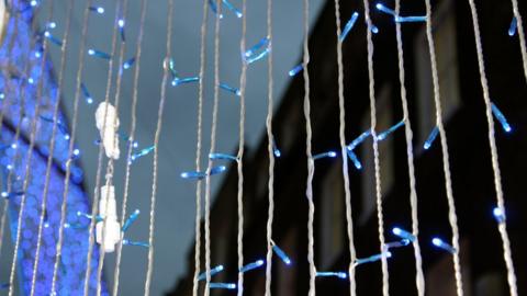 Columns of strings of Christmas lights, which are illuminated blue. A larger blue light installation is illuminated but out of focus in the background.