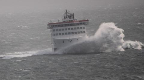 The Manxman ferry, which is a large white ferry, with a large wave crashing across the bow in choppy seas.