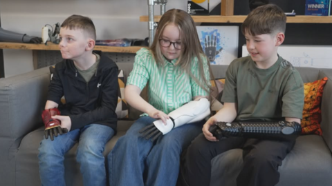 Three children, two boys with a girl in the middle, sitting on a sofa, all aged about nine and wearing their new bionic arms. They are looking at them and feeling what's they're like to wear