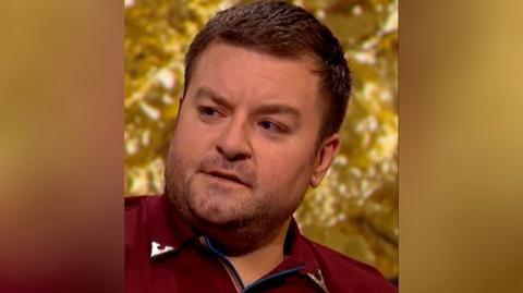 Alex Brooker in front of a gold background