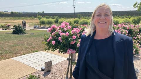 Astrud stands in front of a row of pink rose trees (bushes that are flowering at the top of a trunk). She smiles at the camera and her blonde hair blows slightly in the wind. Behind and to her right is a gravel path with occasional headstones. Beyond that is a grass lawn an more small green trees. She wears a blue suit jacket over a navy round necked shirt.
