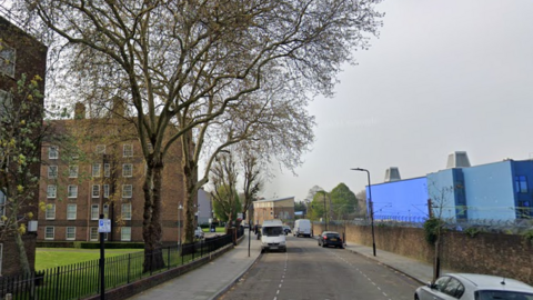 Google StreetView image showing art of Bodney Road. A period block of flats is visible on the left of the road, while a large blue industrial structure can be seen on the right