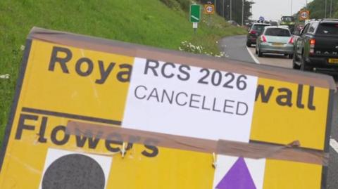 A yellow Royal Cornwall Show road sign has a temporary note stuck on top of it with tape, saying "RCS 2026 Cancelled".