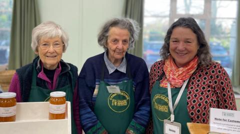 Dorothy, Jean and Christa are standing side-by-side, wearing green aprons with "Cambridgeshire county markets" written on them. There are two jars of marmalade on stands in front of them.