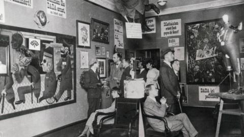 A group of people sit in an art gallery. The paintings and sculptures are all Dada.