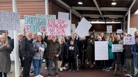 Dozens of people stood outside a large brown building while a handful hold placards with messages to stop adult social care cuts