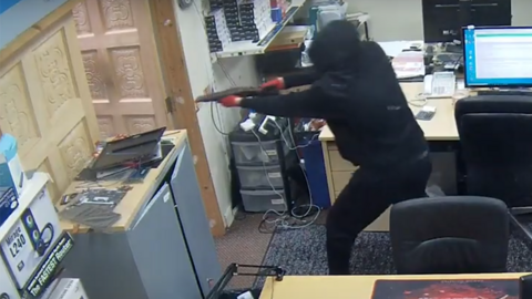 A masked man holding a sawn-off shotgun which is being pointed towards an open door. He is wearing a black hoodie with the hood up, and red gloves. He is standing in what looks to be an office with desks and drawers inside