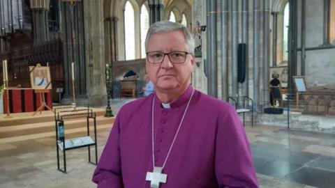 The Bishop of Salisbury stands in Salisbury Cathedral in purple clergy garments with a large cross round his neck.