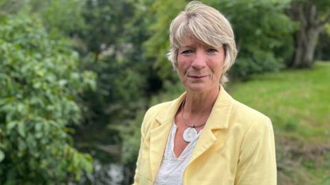 Pippa Heylings looks directly at the camera, smiling slightly. She is standing on a riverbank with a body of water behind her. She is wearing a pale yellow jacket and white top, with a silver necklace. She has short blonde hair. 