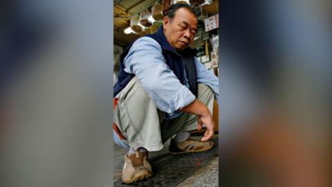 A man sits in the position of an 'Asian squat'. He's wearing a blue shirt and green trousers.