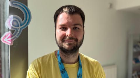 Pierce is a young man with short, brown hair and facial hair. He is smiling at the camera with his mouth closed. He is wearing a yellow t-shirt and a blue lanyard with Eastside Partnership logo on it. Behind him is a blank wall.