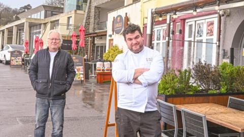 Larry Gibbons and Tom Robinson stand outside their cafes which are next door to each other. Tom is wearing a white chef's jacket and has his arms folded. Larry is wearing black jeans and a jacket and has his hands in his pockets.