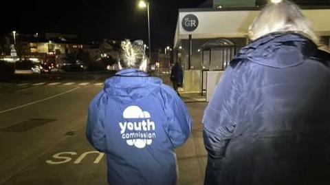 A back view of two youth support workers walking in the dark in Guernsey. The two women are wearing navy jackets, the one on the left has a logo reading Youth Commission.