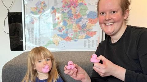 A woman with short blonde hair sits next to a child with long blonde hair. They woman is holding two small pink rubber ducks and the child has one in her mouth. Behind them is a map of the UK with pink dots all over it. 