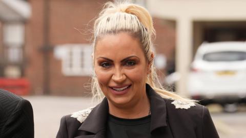 Jodie Marsh has blonde hair in a high ponytail. She is wearing a black top and a black blazer with silver detail on the shoulders. Behind her is a building and a parked white car.