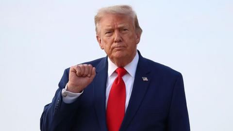 Donald Trump holds up his right fist while wearing a white shirt and red tie.