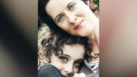 Anne-Marie Cockburn, a woman with dark hair and grey eyes and wearing a chunky necklace, smiles with her daughter Martha, a young girl with the same colour eyes and brown curly hair.