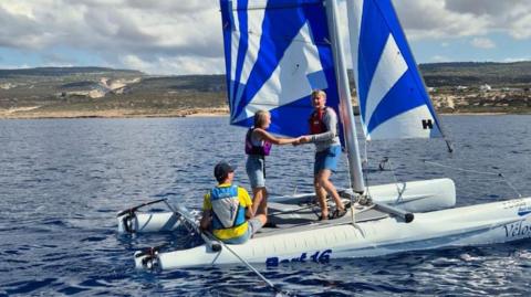 A man and a woman in red life jackets dance on board a small catamaran with a blue and white sail while a man in a yellow top steers the vessel. 