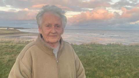 Celia Henry stands with the estuary of the Wansbeck from Cambois beach behind her. She is an elderly woman wearing a brown fleece. The sky is full of pink and grey clouds.