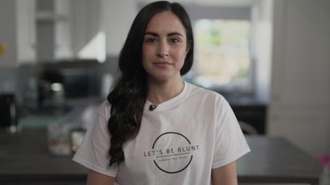 Leanne Lucas, who has long dark hair, smiles at the camera while wearing a white t-shirt with the logo 'Let's Be Blunt - Remove the Point'