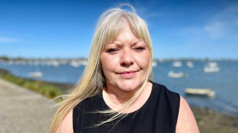 A head and shoulders image of Lisa Morris standing on a footpath next to a waterway, possibly the sea. She has long blond hair which is being blown by the wind.