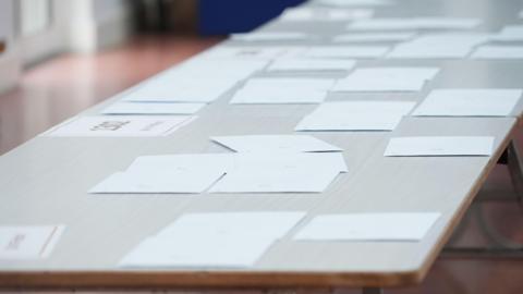 Several results envelopes on a table in a school classroom. They are white with stickers on them.