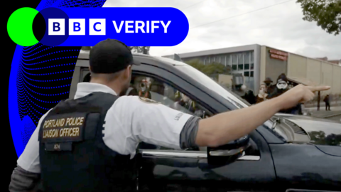Man wearing vest which says "Portland Police Liaison Officer" is speaking into an open car window and pointing 