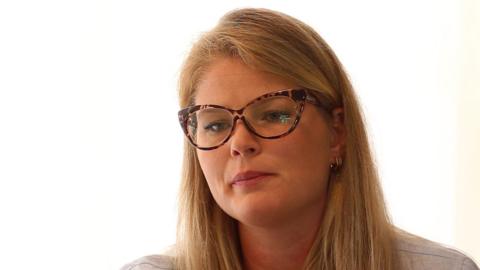 Headshot of a woman with long straight blonde hair. She is looking pensive and is wearing tortoiseshell glasses. She is wearing a pale beige top. 