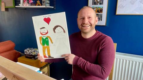 A man in a maroon jumper sits on a dining table chair holding a white canvas. On it is a crude painting of a red love heart, a man in a green and yellow suit and a woman in a white dress. The man holding the artwork has very short hair and a trimmed beard. He is laughing. Behind him, there are a couple of boxes, a white shelf, a white radiator, a couple of pictures and a blue wall.