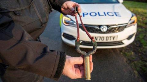 A police officer stands in front of a patrol car and holds a catapult.