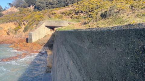 Concrete sea defences with a fun emplacement to the left and rocks behind.