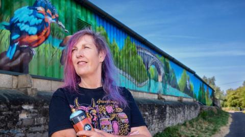 A woman stands in front of a colourful mural and holds a spray can. She has purple hair and glances off to the side