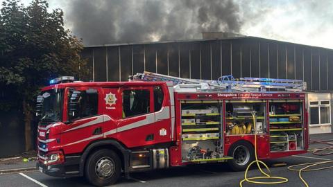 A fire engine is outside a factory warehouse building with large clouds of thick, black smoke above it. There is a yellow hose leading from the engine.