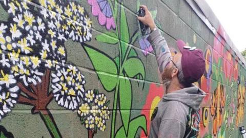 A street artist wearing a purple baseball cap and grey hoodie top spray paints a flower onto a brick wall.