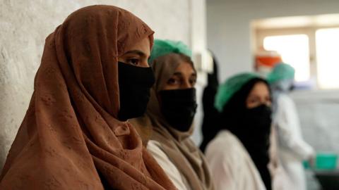 Three women wearing masks and hijabs, sit in a line, as a woman makes jam in the background