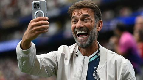 Jurgen Klopp smiling while holding a mobile phone in front of his face