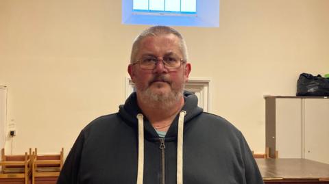 A middle aged man with glasses and grayish brown hair. He is wearing a hoodie and standing in front of a off-white wall. 