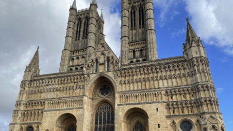 The photo has been taken from the ground and pans up to Lincoln Cathedral. On the left hand side the sky is grey and cloudy, but on the right hand side it is blue. 
