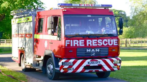 A red fire engine from Devon and Somerset Fire Service on a road surrounded by green lawns on a sunny day. A knitted firefighter toy is on the dashboard along with another cuddly toy.