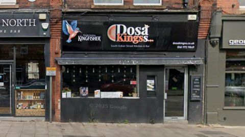 The front of the Dosa Kingss restaurant on the high street, including a black sign above the front window and entrance. 