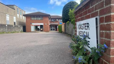 The sign outside Caister Medical Centre - which has since been renamed Caister Health Centre. The picture shows the signage with flowering weeds growing nearby. The section of red brick wall leads to a wooden fence, which is partially covered in ivy, with a tree growing above. The Caister Health Centre buildings can be seen to the centre left of the image; a 1980s two-storey building, built of red brick with hipped tiled roofs. Windows and air conditioning units can be seen in the image.